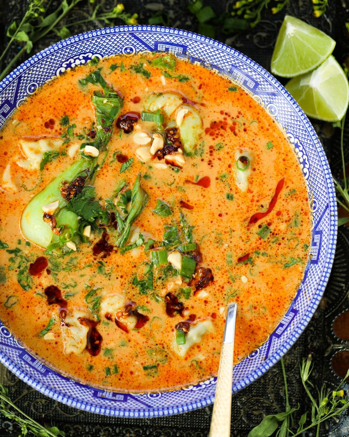 A bowl of creamy orange soup topped with dumplings, greens, herbs, chili oil, and chopped peanuts, with lime wedges on the side.