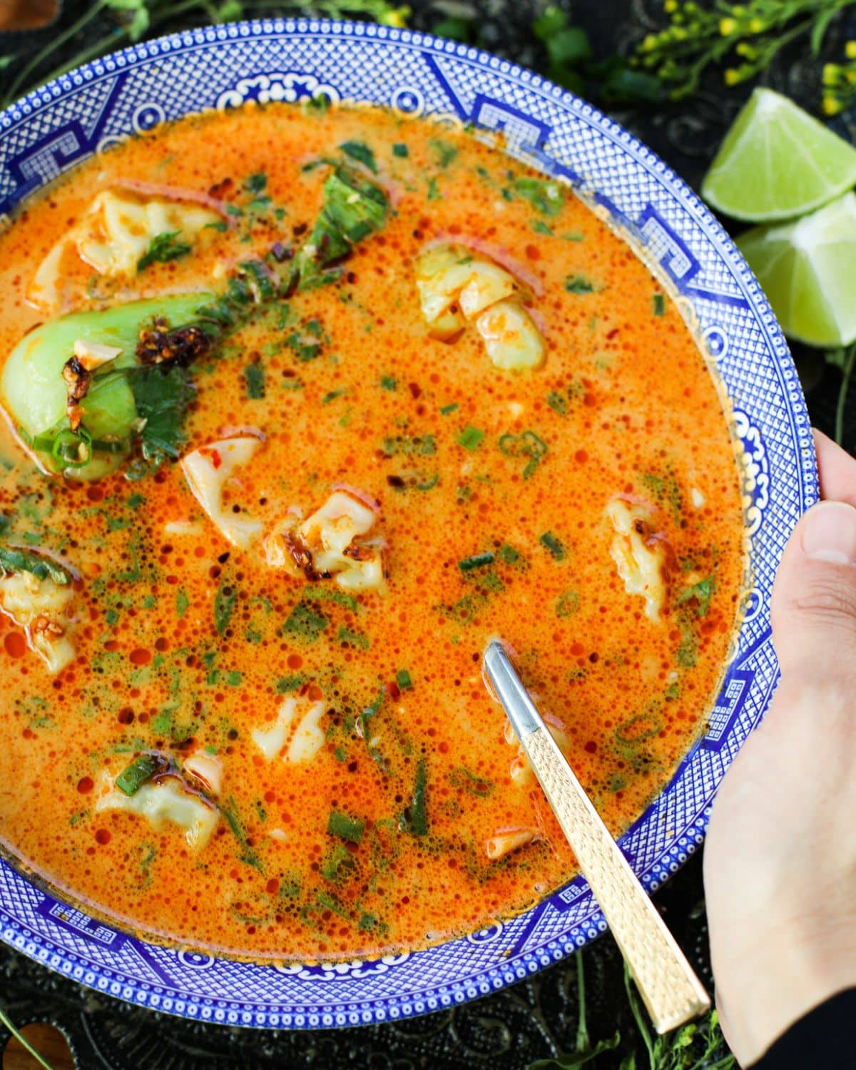 A bowl of orange-red soup with dumplings, greens, and herbs, served with a spoon in a blue patterned bowl; lime wedges are visible on the side.