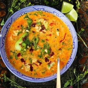 A bowl of creamy orange soup garnished with fresh herbs, chili oil, and chopped peanuts, with lime wedges on the side. A spoon rests in the bowl.