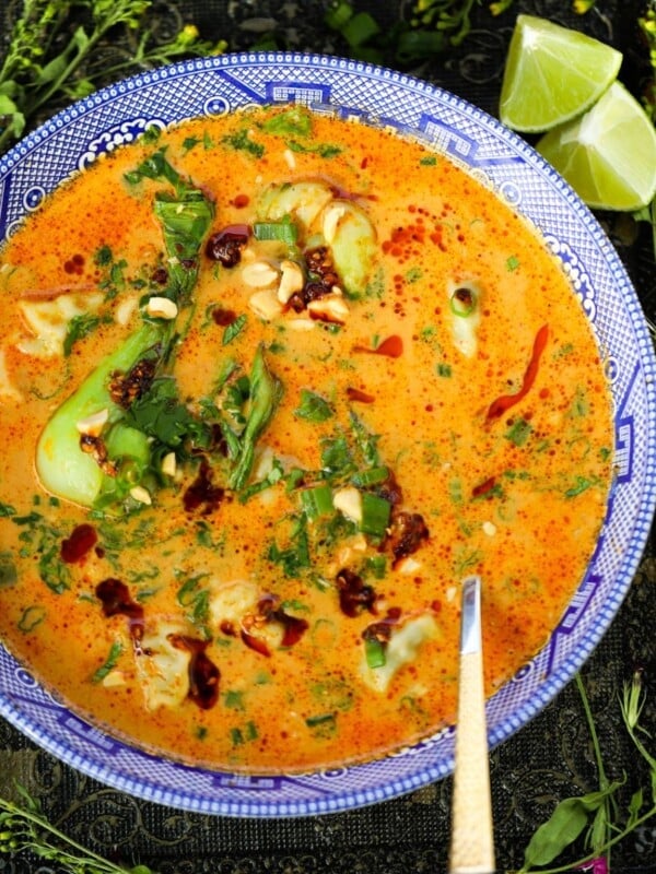 A bowl of creamy orange soup garnished with fresh herbs, chili oil, and chopped peanuts, with lime wedges on the side. A spoon rests in the bowl.