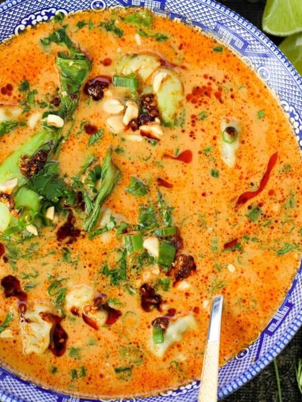 A bowl of spicy orange soup with bok choy, peanuts, herbs, chili oil, and green onions, served with a spoon and lime wedges on the side.