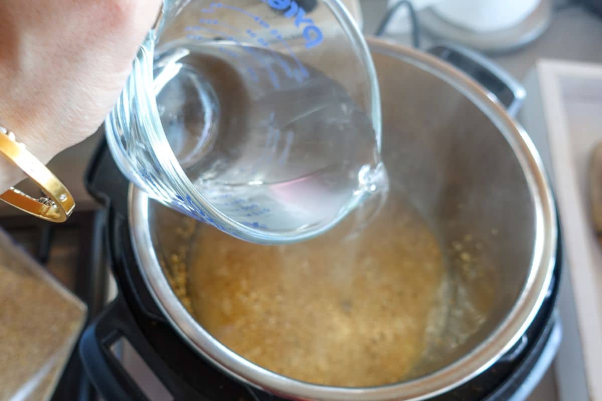 A hand pours water from a measuring cup into an Instant Pot containing cracked wheat.