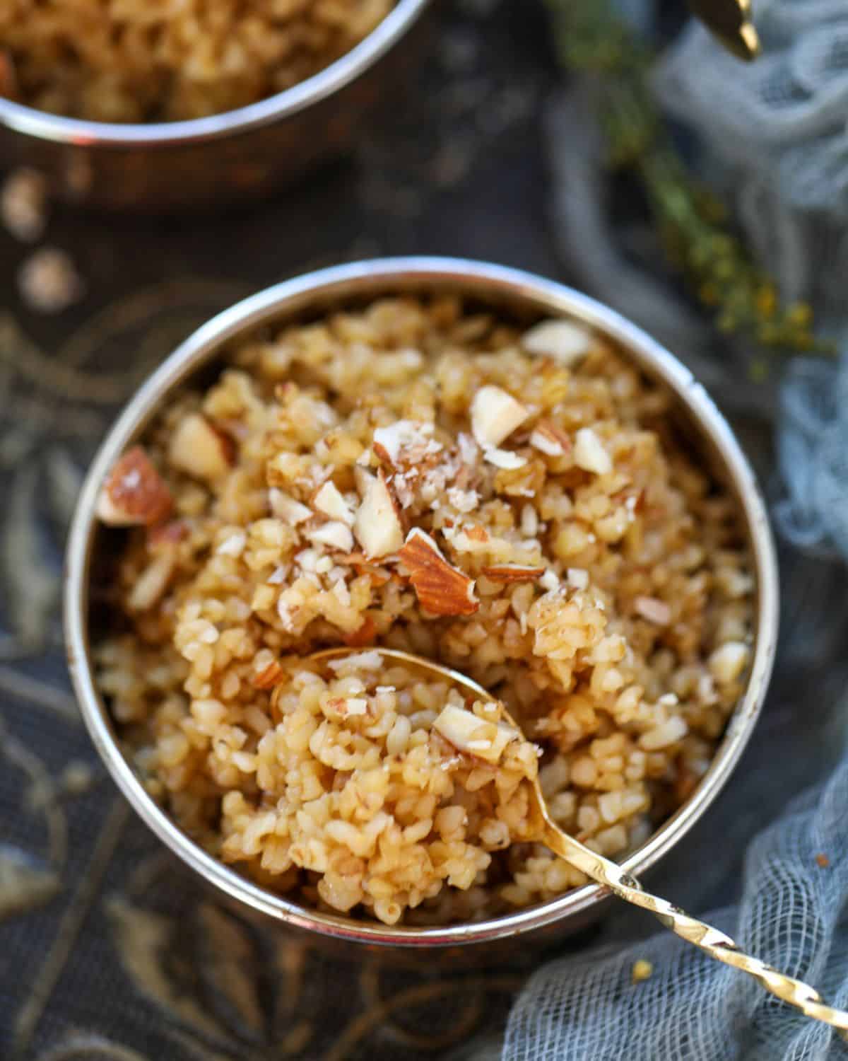 A bowl of cooked sweet daliya topped with chopped almonds, with a spoon resting in the bowl, placed on a dark patterned cloth.