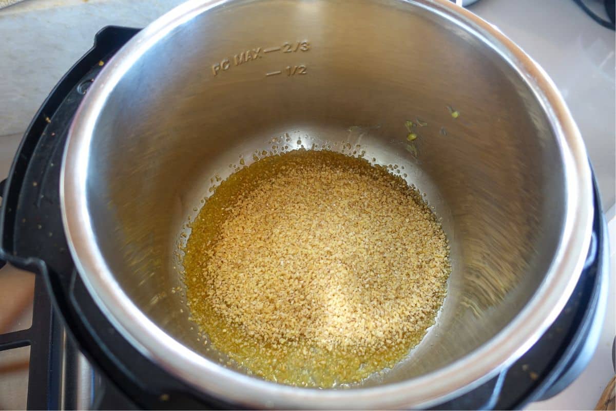 Cracked wheat being toasted in ghee inside a stainless steel Instant Pot on a kitchen countertop.