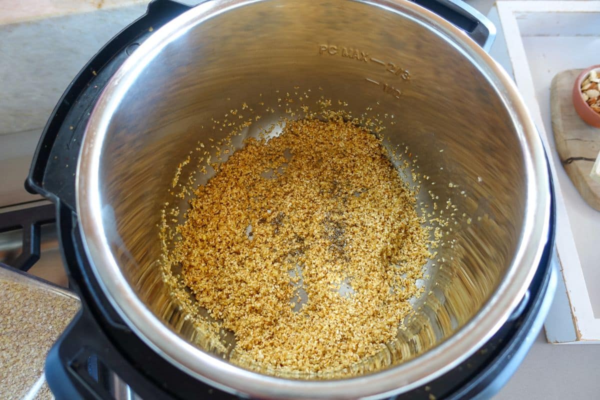 Close-up of an Instant Pot interior with saut&eacute;ed cracked wheat partially browned and cardamom. A wooden cutting board with nuts is visible in the background.