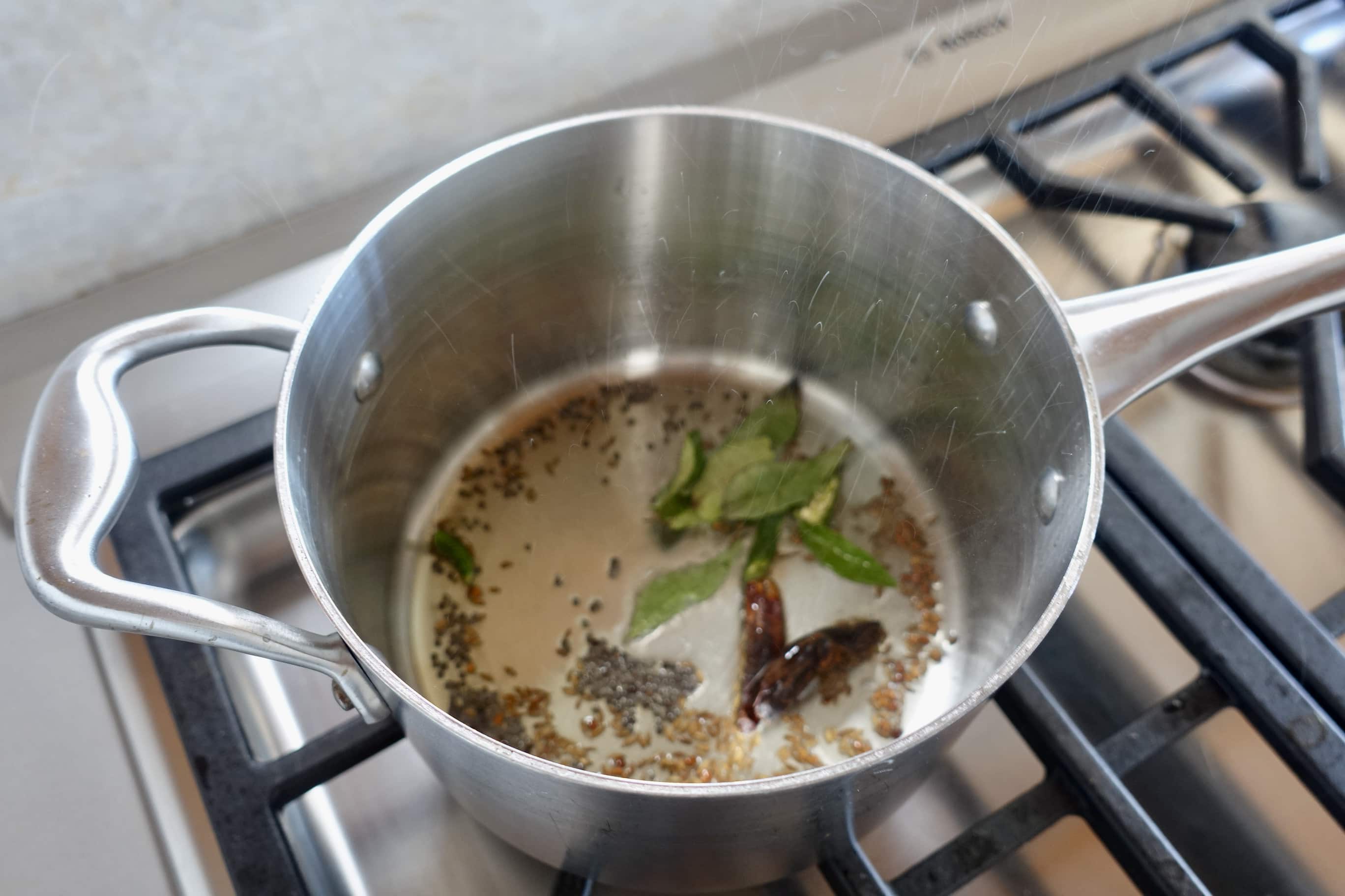 A stainless steel pot on a stovetop with spices, dried chilies, and curry leaves being saut&eacute;ed in oil.