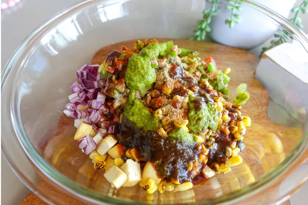 A glass bowl with chopped red onions, corn, diced tomatoes, sauces, spices, and green chutney on a wooden board.