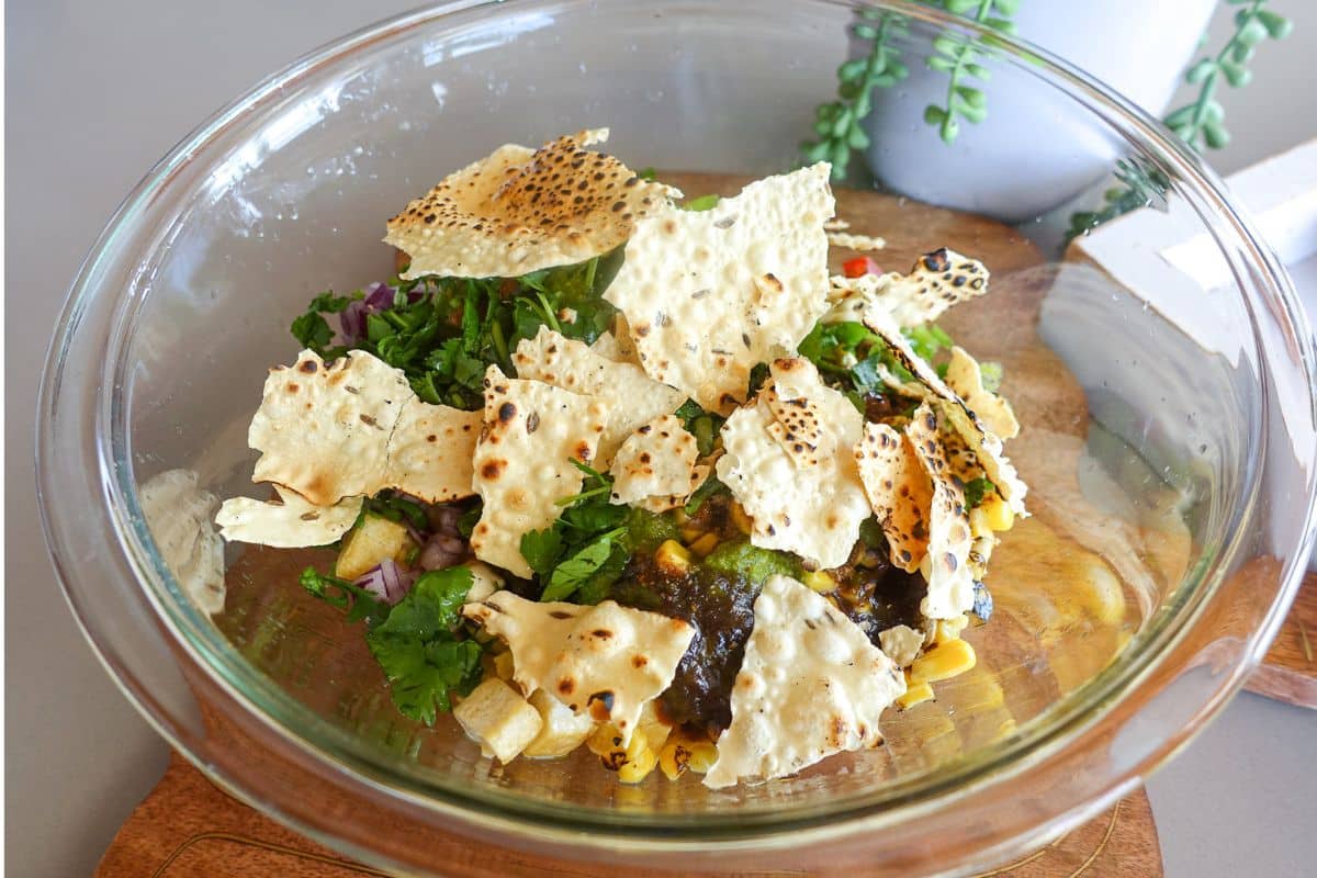 A glass bowl containing a mixture of vegetables, herbs, and broken pieces of crispy flatbread, placed on a wooden board.