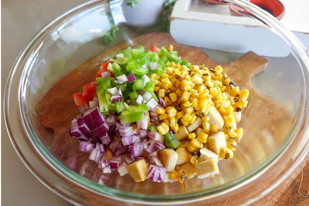 A glass bowl contains chopped red onions, green bell peppers, tomatoes, and roasted corn on a wooden board.