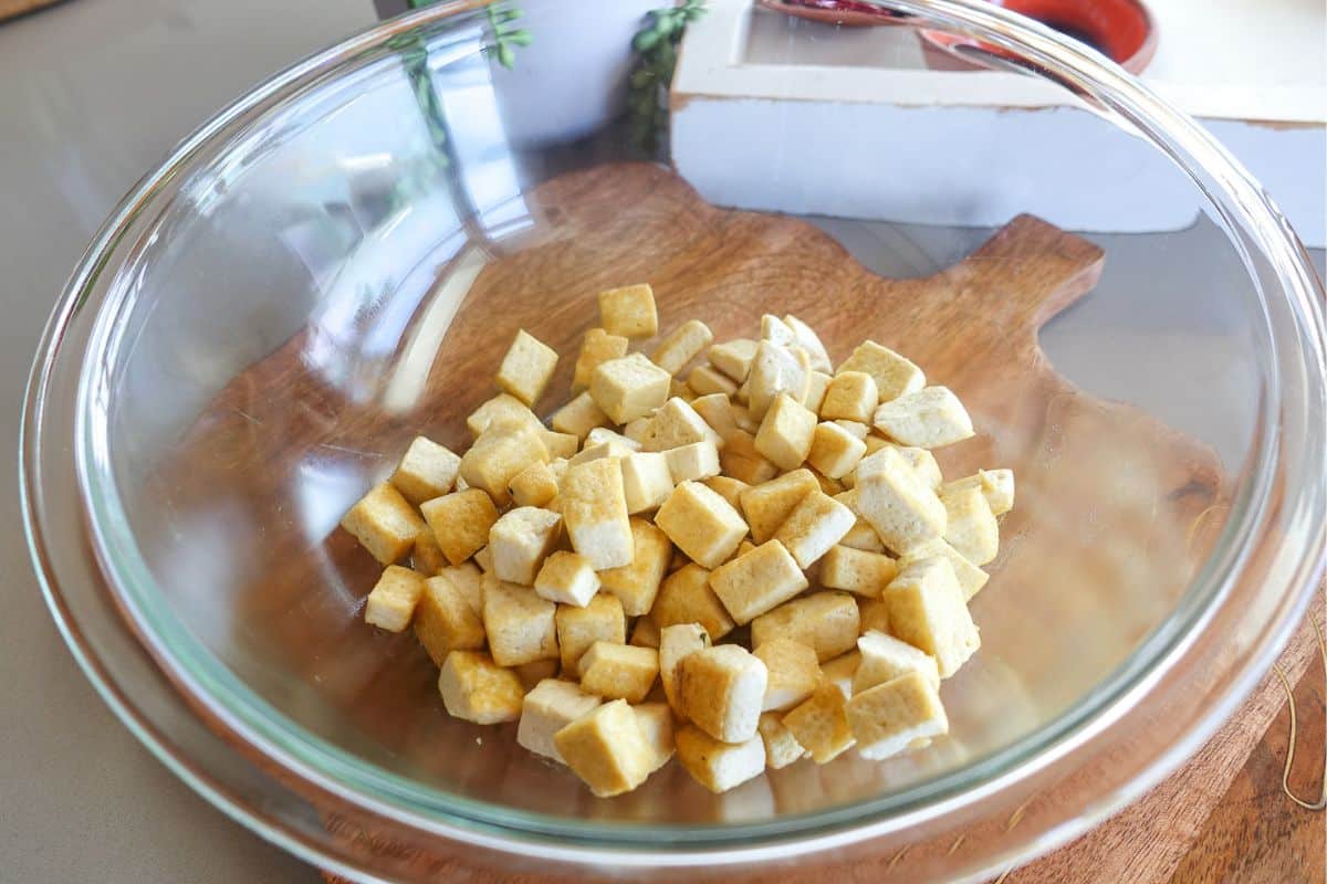 A clear glass bowl containing cubed pieces of tofu sits on a wooden cutting board.