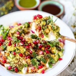 A plate of high protein dahi chana chaat topped with sev, chickpeas, pomegranate seeds, chopped herbs, and sauces, with a spoonful being lifted.