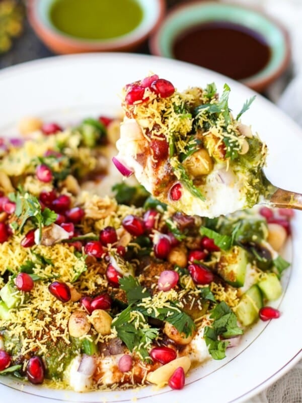 A plate of high protein dahi chana chaat topped with sev, chickpeas, pomegranate seeds, chopped herbs, and sauces, with a spoonful being lifted.