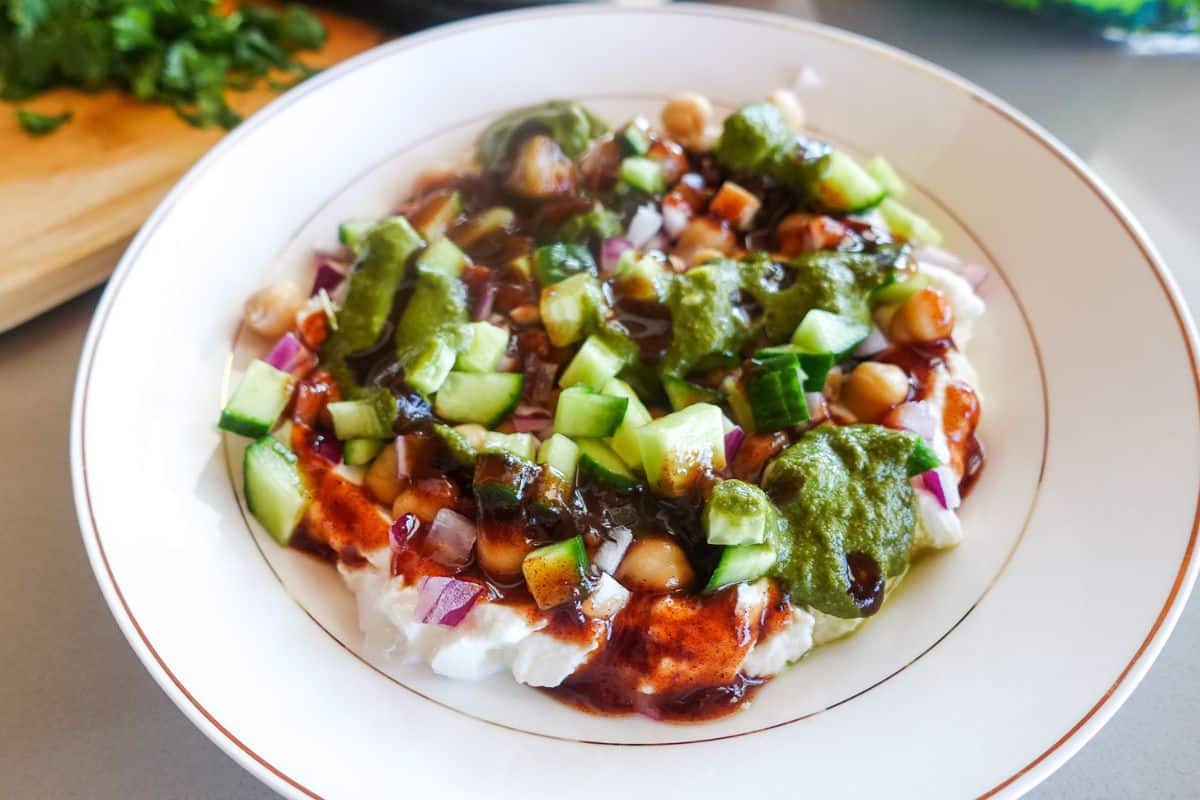 A bowl of dahi chaat topped with chopped cucumbers, onions, chickpeas, green chutney, and tamarind chutney.
