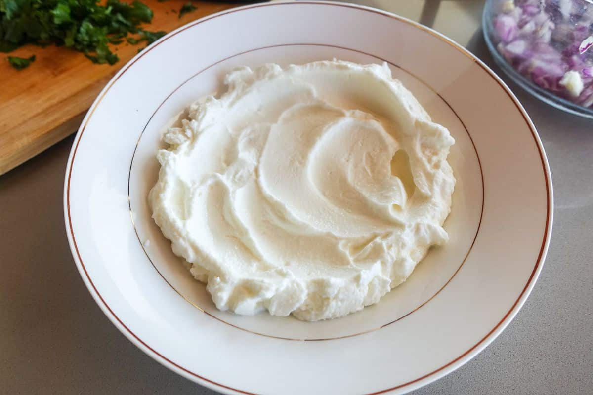 A white bowl filled with smooth, Greek yogurt on a countertop, with chopped herbs and onions visible in the background.