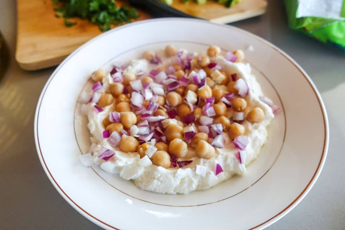 A white plate with a creamy yogurt base topped with chickpeas and chopped red onions, placed on a countertop near a cutting board with herbs.