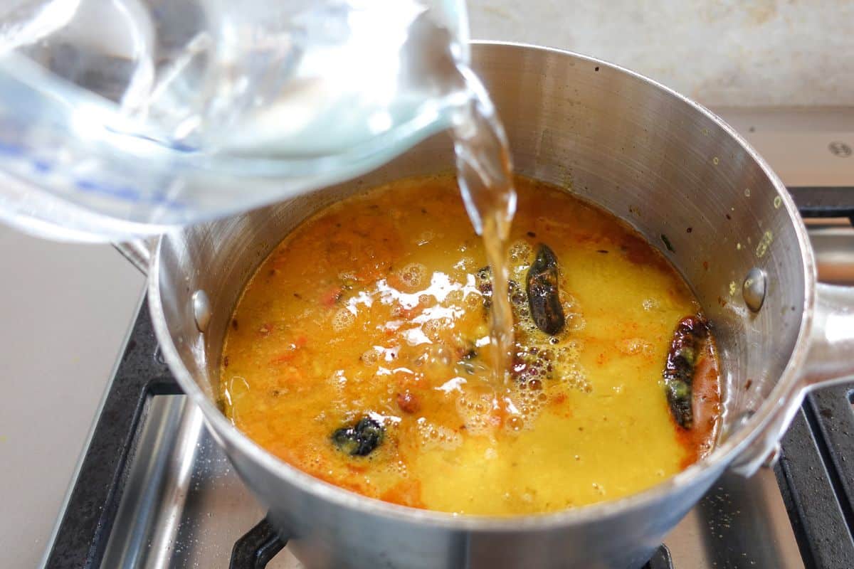 Water is being poured into a pot of simmering toor dal with spices, on a stovetop.