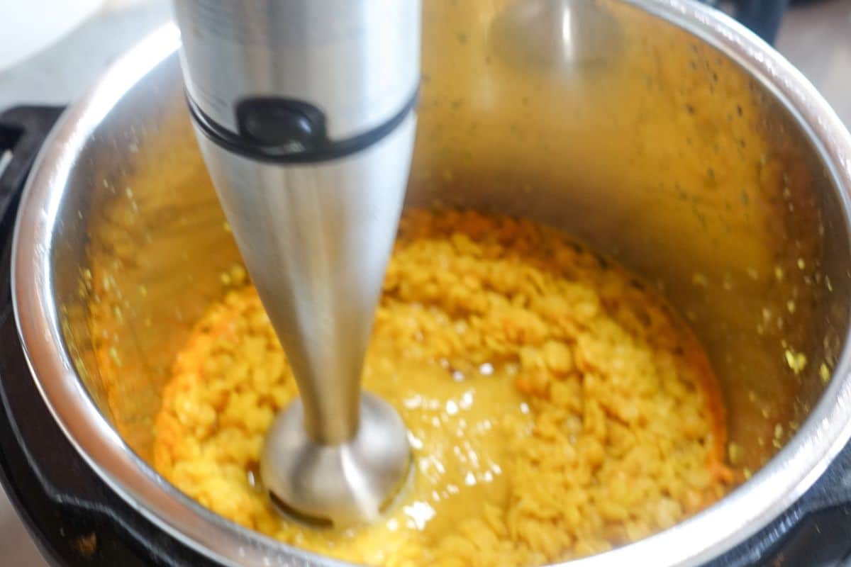 An immersion blender is being used to blend cooked yellow lentils in a stainless steel pot.