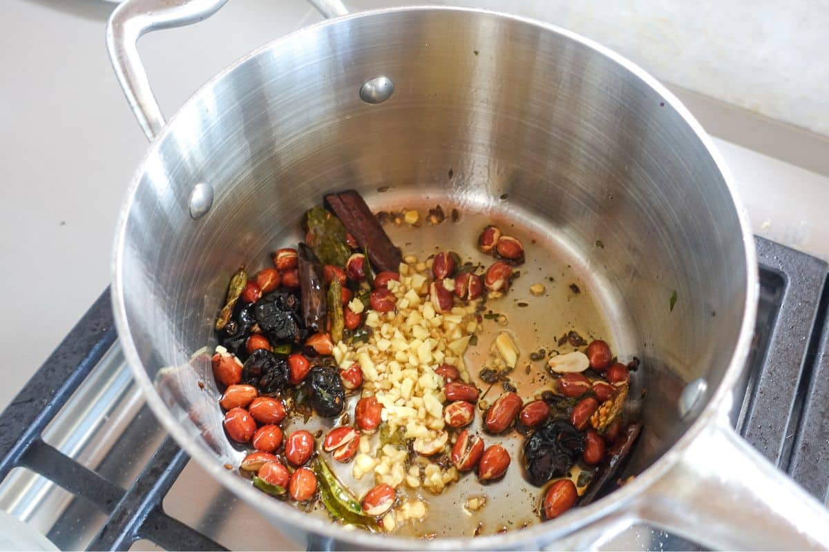 A stainless steel pot on a stove contains peanuts, chopped ginger, dried chilies, whole spices, and oil being saut&eacute;ed.