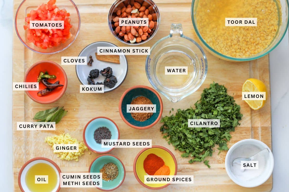 A wooden board displays labeled ingredients for dal including tomatoes, peanuts, toor dal, spices, herbs, water, oil, ginger, chilies, lemon, and salt in small bowls.