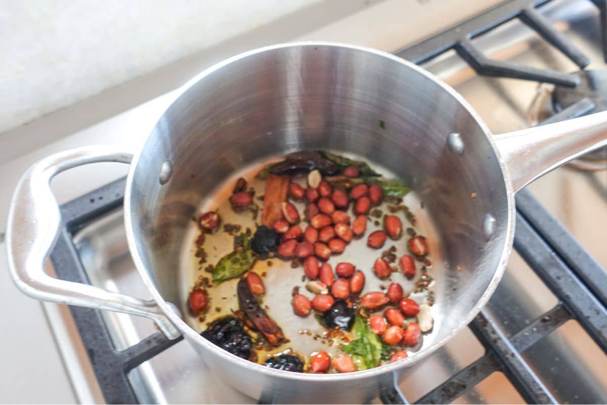 A stainless steel pot on a stove contains peanuts, dried chilies, curry leaves, and spices being tempered in oil.