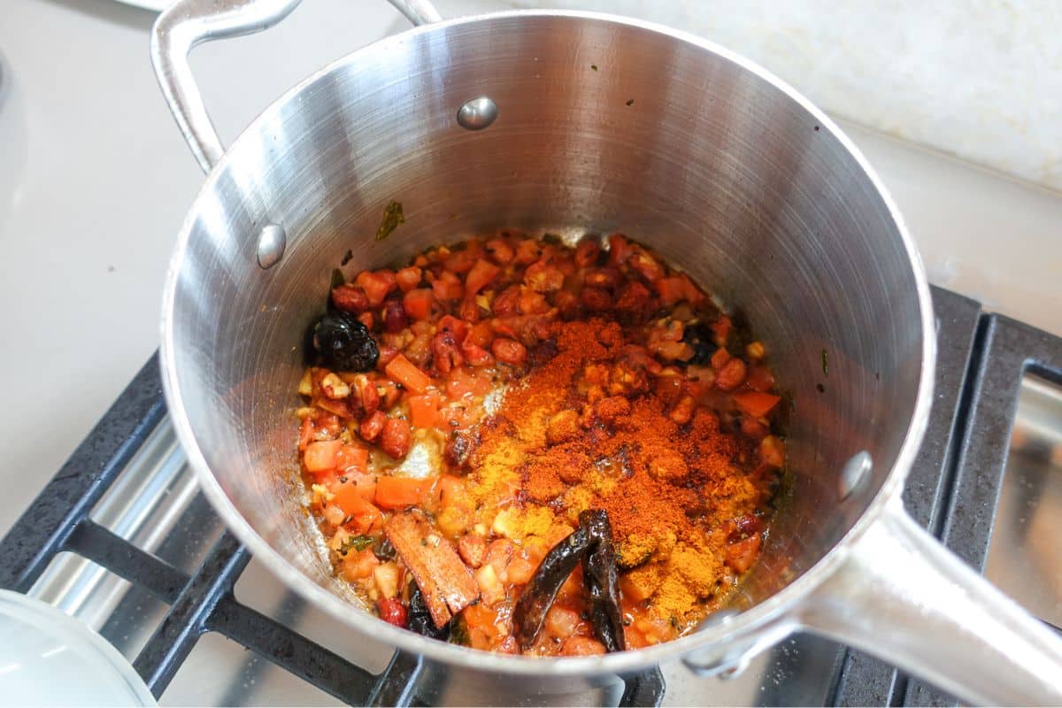 Chopped tomatoes, whole dried chilies, and ground spices cooking in a stainless steel pot on a stovetop.