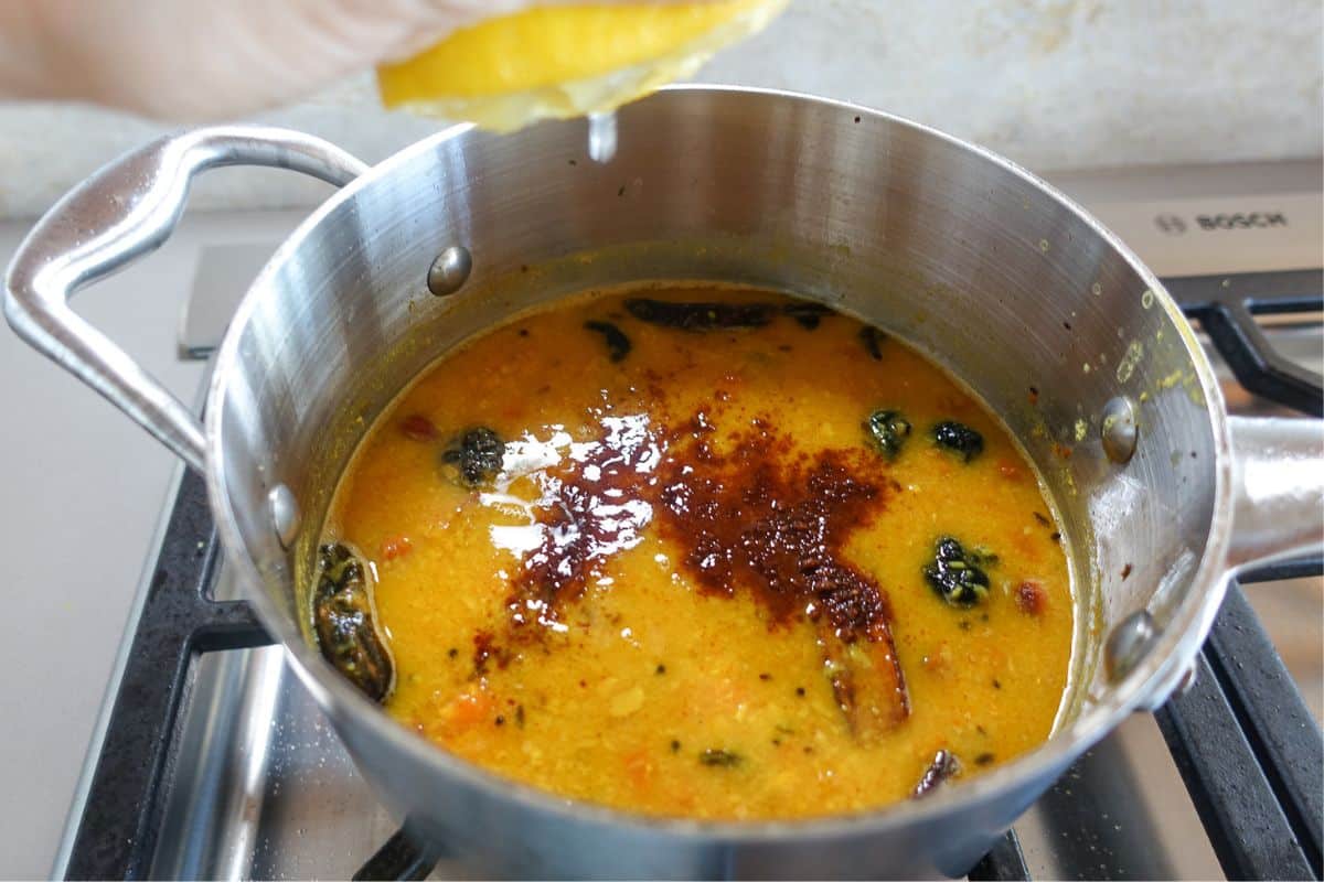 A hand squeezes a lemon over a pot of yellow dal with jaggery cooking on a stovetop.