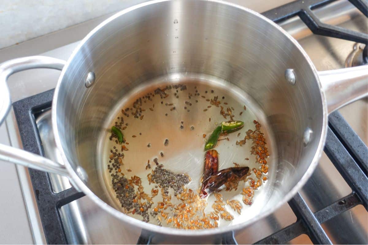A stainless steel pot on a stove contains oil with cumin seeds, mustard seeds, dried red chili, and sliced green chili being tempered.