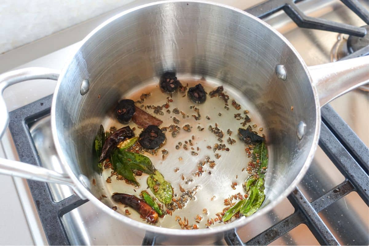 A stainless steel pot on a gas stove with oil, dried chilies, curry leaves, kokum, and spices being saut&eacute;ed.