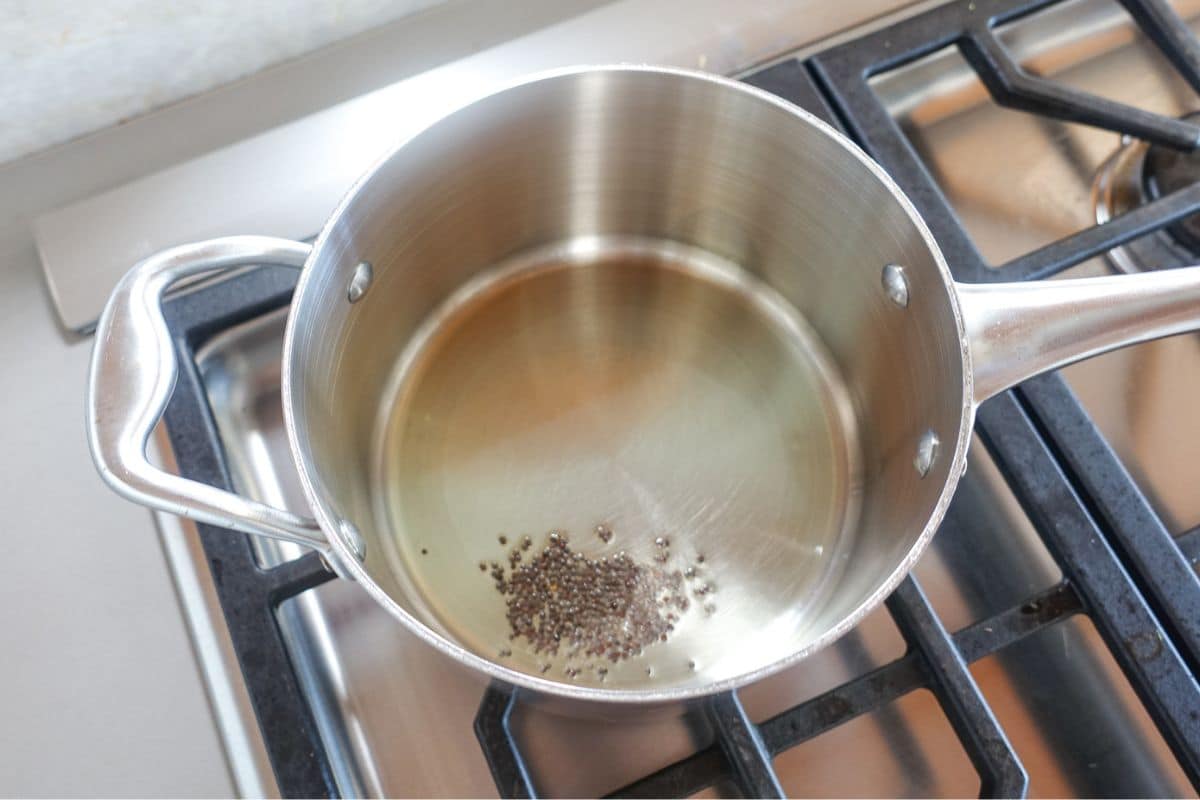 A stainless steel pot with hot oil and a small amount of mustard seeds sits on a gas stove burner.