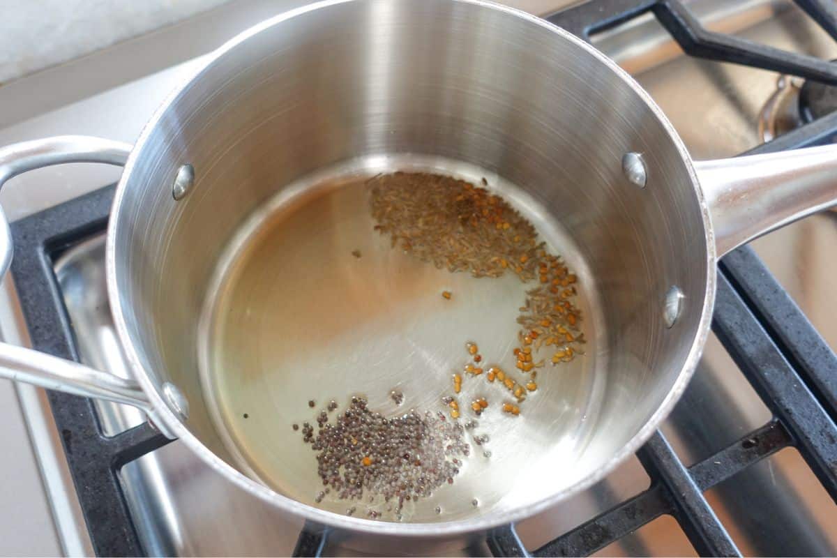 A stainless steel pot on a stove contains oil with methi seeds, cumin seeds, and mustard seeds starting to sizzle.