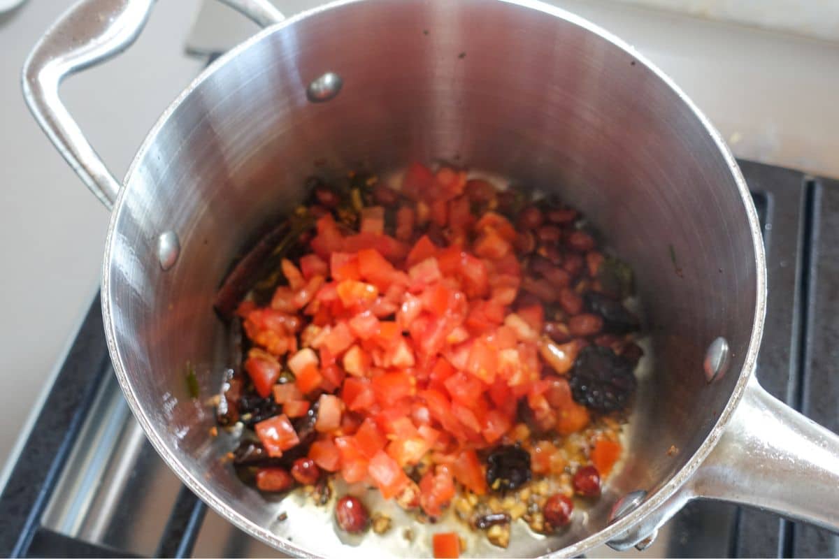 Chopped tomatoes and mixed ingredients including peanuts are cooking in a stainless steel pot on a stovetop.