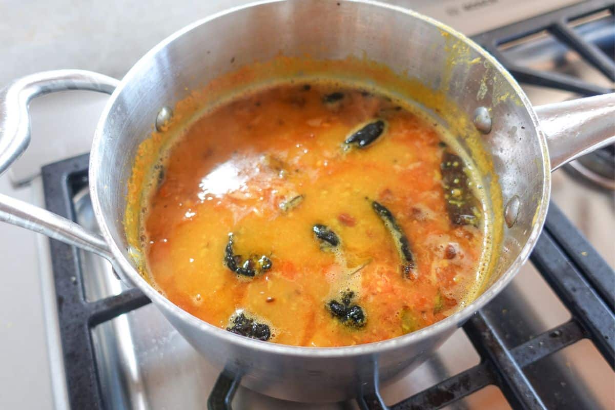 A stainless steel pot on a stovetop contains simmering yellow-orange toor dal with visible spices and whole dried chilies.