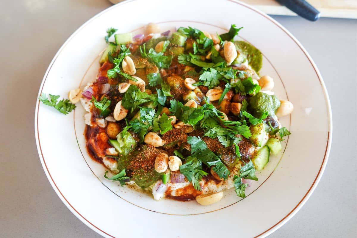 A plate of Indian chaat topped with chopped onions, cucumbers, peanuts, green chutney, spices, and fresh cilantro on a white plate.