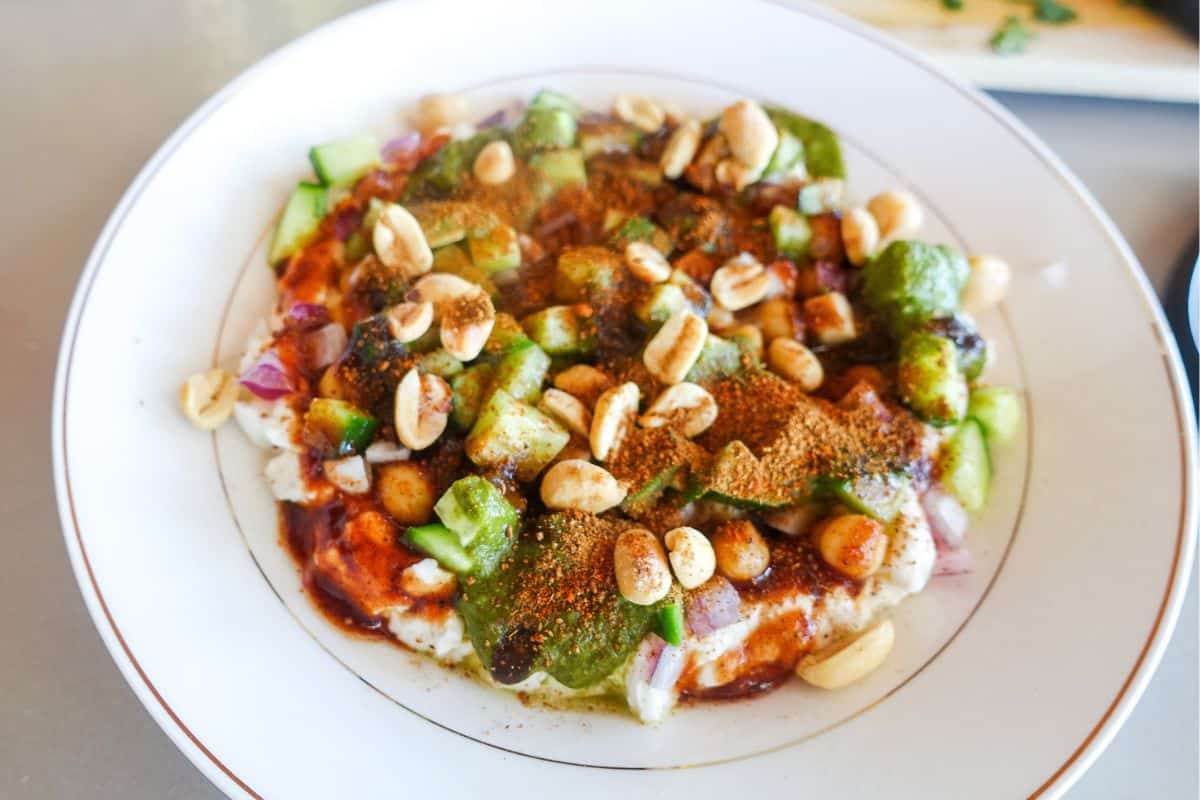 A white plate containing dahi chaat topped with chopped vegetables, peanuts, yogurt, green chutney, tamarind chutney, and spices.