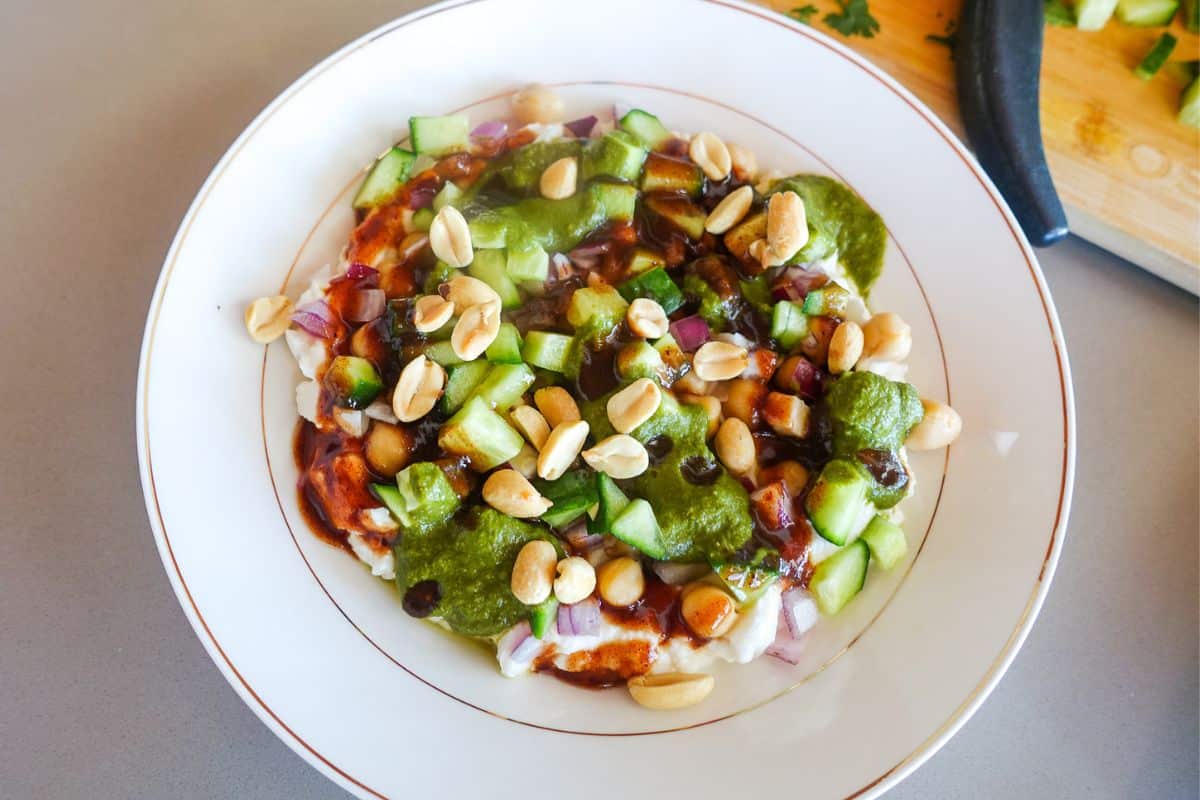 A plate of dahi puri topped with chopped cucumber, onions, peanuts, green chutney, and tamarind chutney, served on a white dish.
