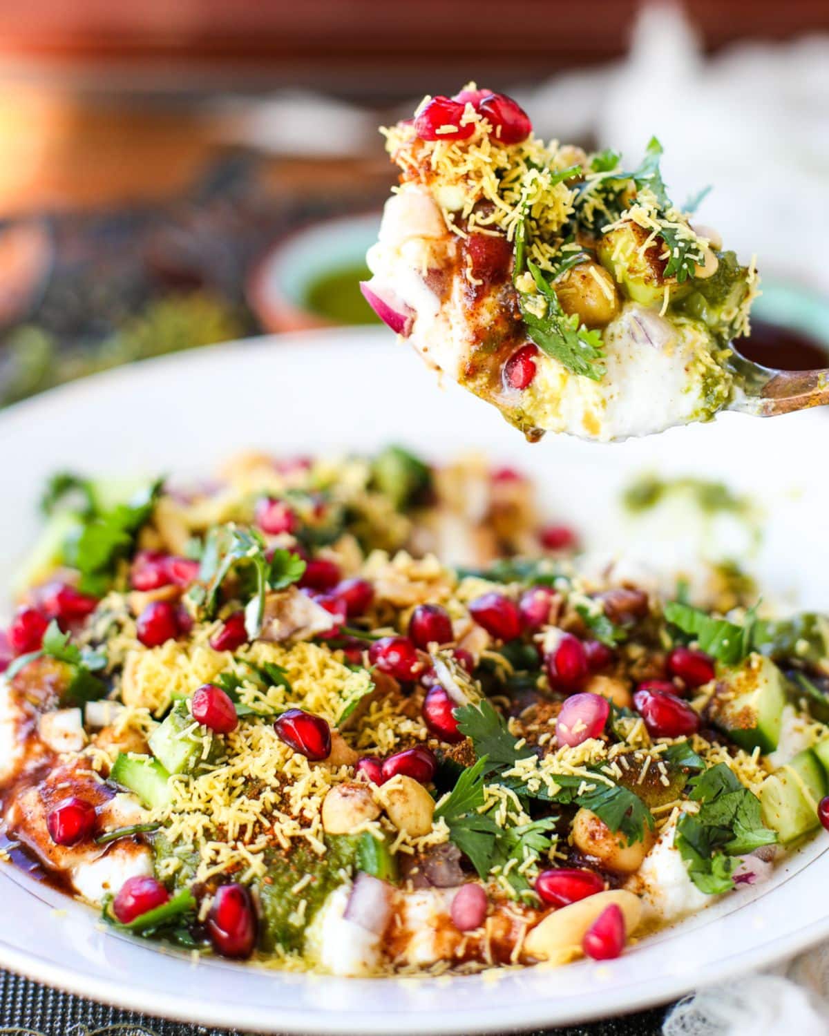 A close-up of dahi puri, an Indian street food dish, topped with sev, pomegranate seeds, cilantro, onions, and chutneys on a white plate, with a spoon lifting a portion.