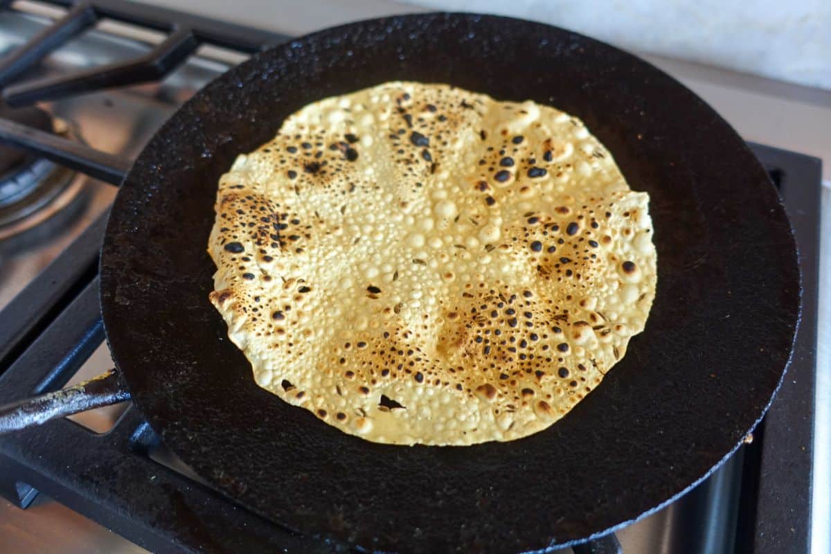 A flat, round papadum with browned spots cooks on a black skillet over a stovetop burner.