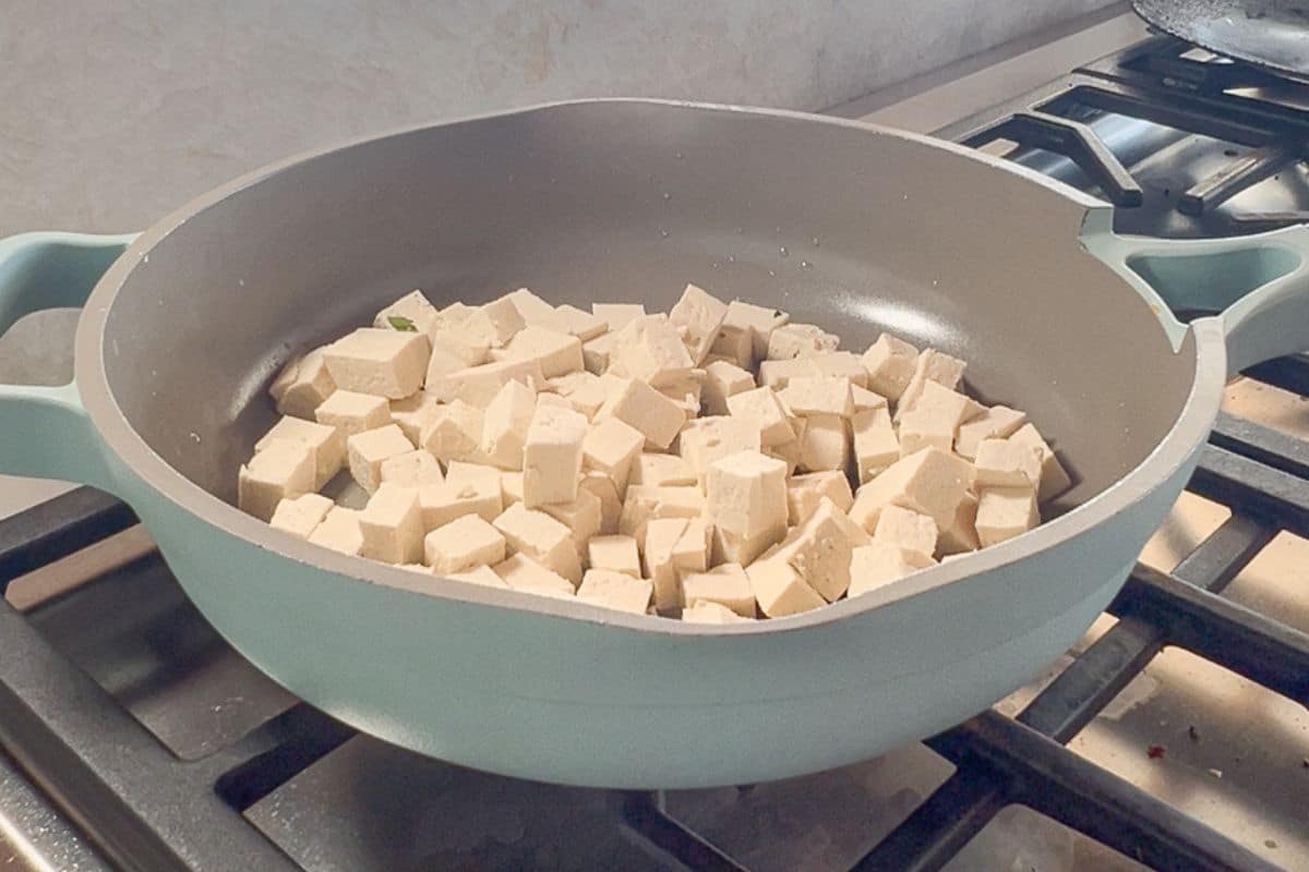 Cubed tofu pieces are being cooked in a light blue nonstick skillet on a gas stove.