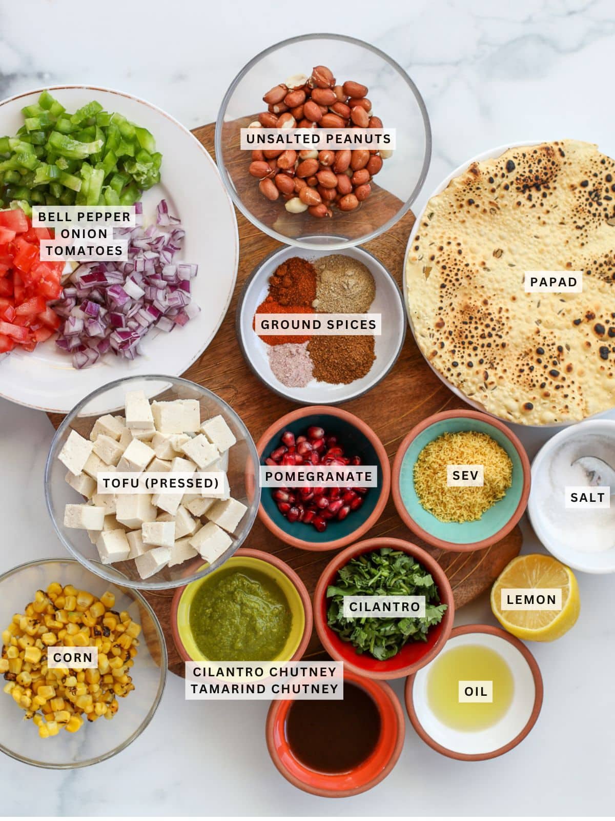 Top view of ingredients for a dish, including chopped bell pepper, onion, tomatoes, tofu, corn, papad, ground spices, chutneys, sev, pomegranate seeds, cilantro, lemon, salt, oil, and peanuts.