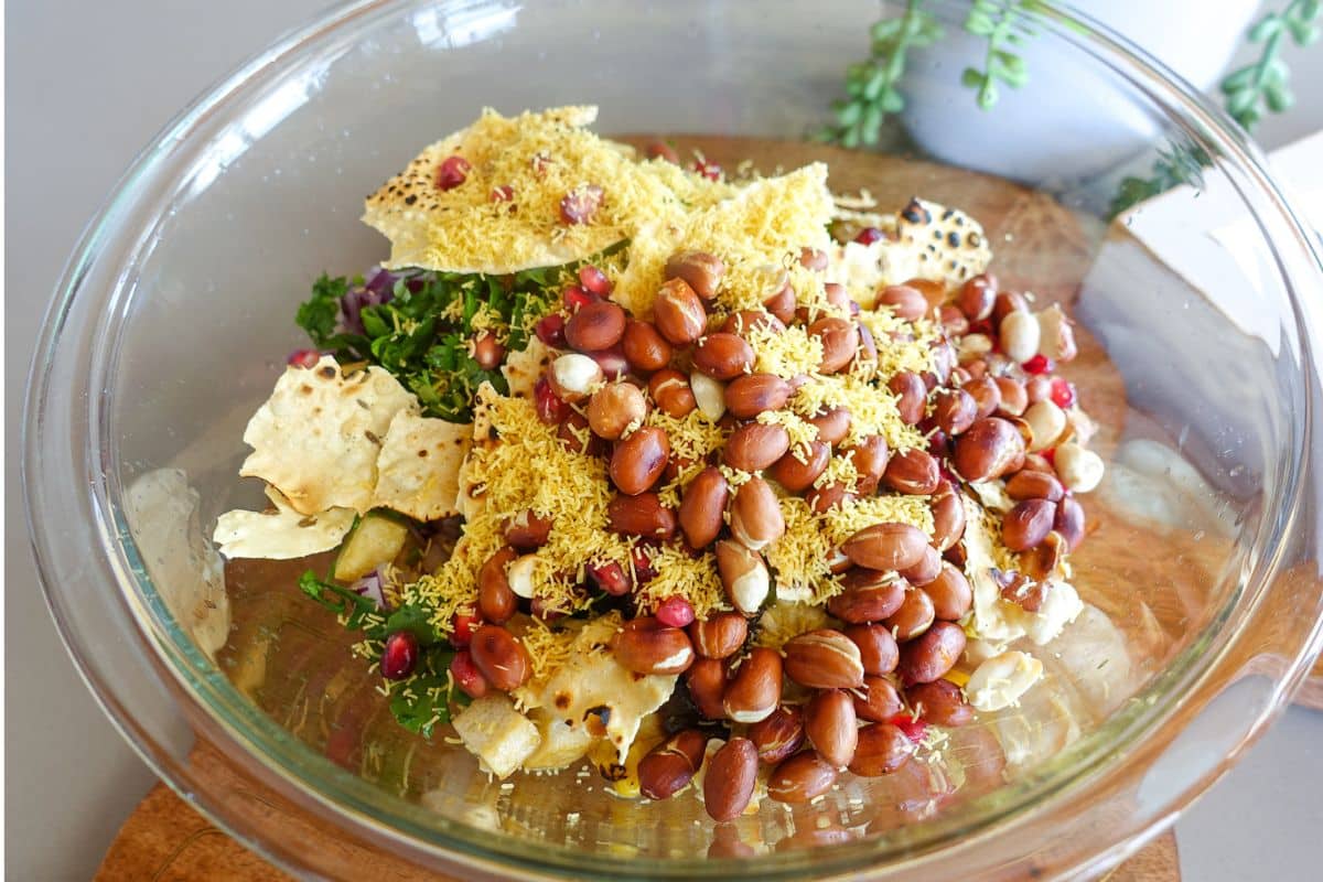 A glass bowl filled with peanuts, sev, pomegranate seeds, crushed papad, and chopped greens, placed on a wooden surface.
