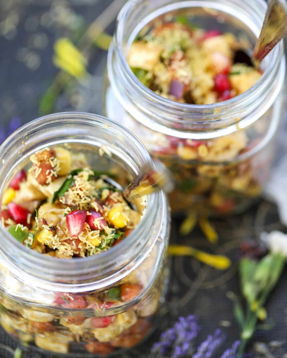 Two glass jars of tofu peanut bhel filled with a colorful mixed fruit and vegetable salad, tofu, garnished with sev and herbs, placed on a dark surface with scattered flowers.
