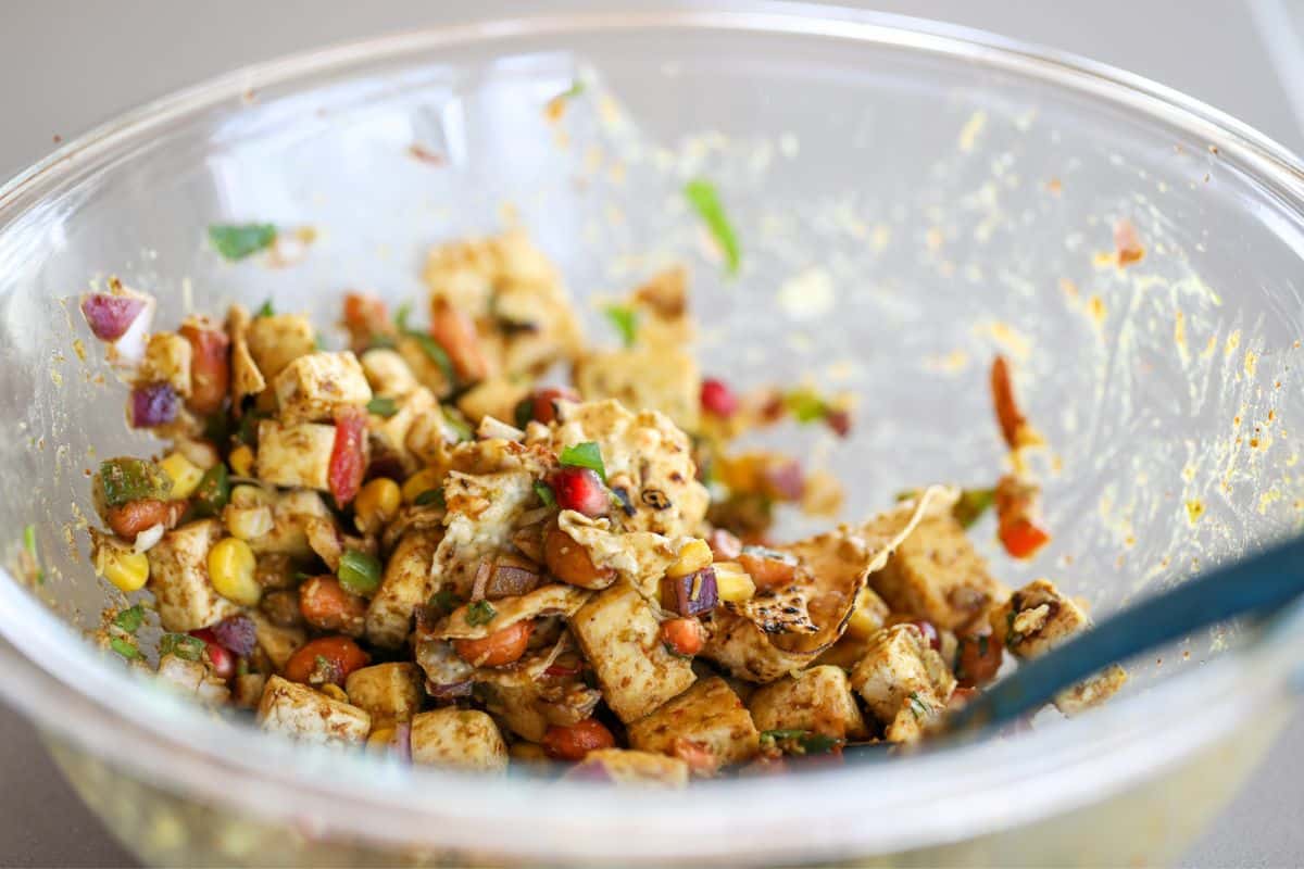 A glass bowl containing a mixed tofu peanut bhel with tofu, corn, diced red onions, peppers, herbs, and seasoning, with a blue utensil on the side.