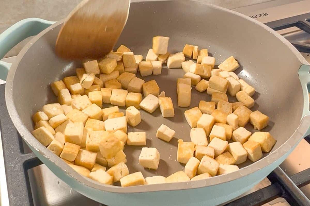 Cubed tofu pieces are being stirred with a wooden spatula in a frying pan on a stovetop.