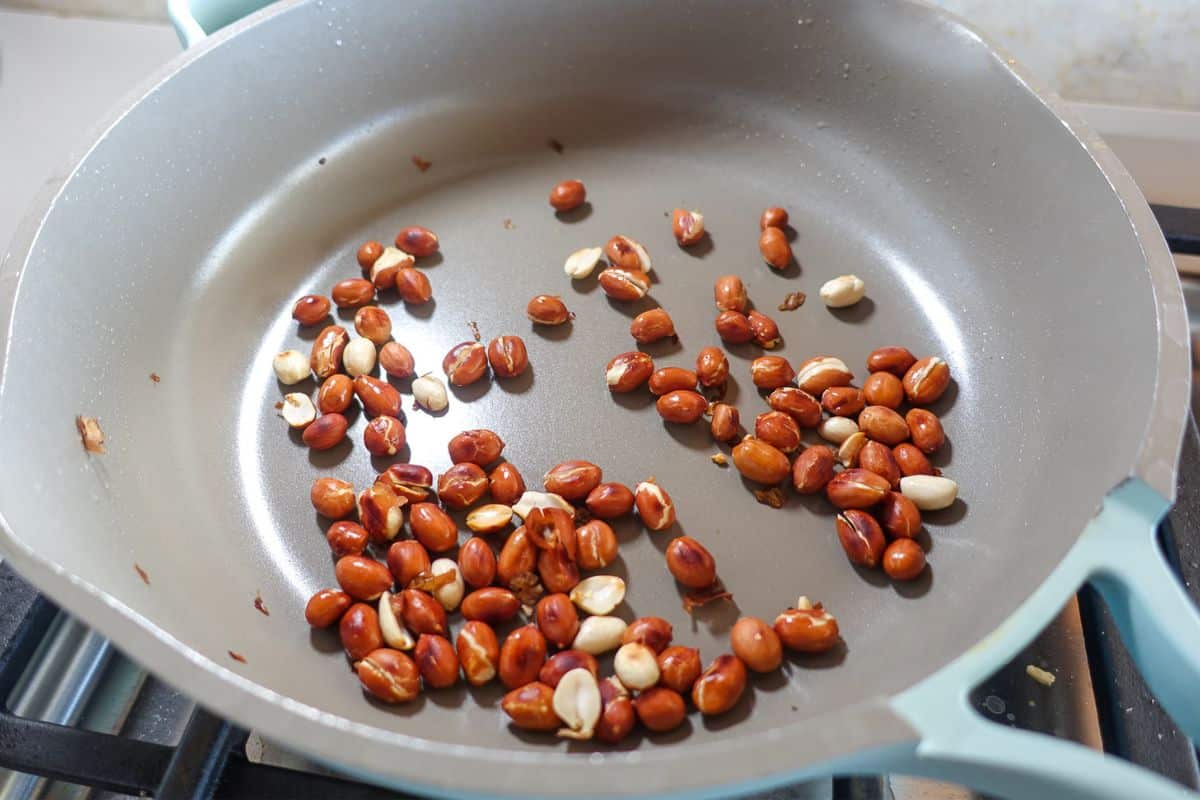 Whole peanuts in their skins are being dry roasted in a non-stick frying pan on a stovetop.