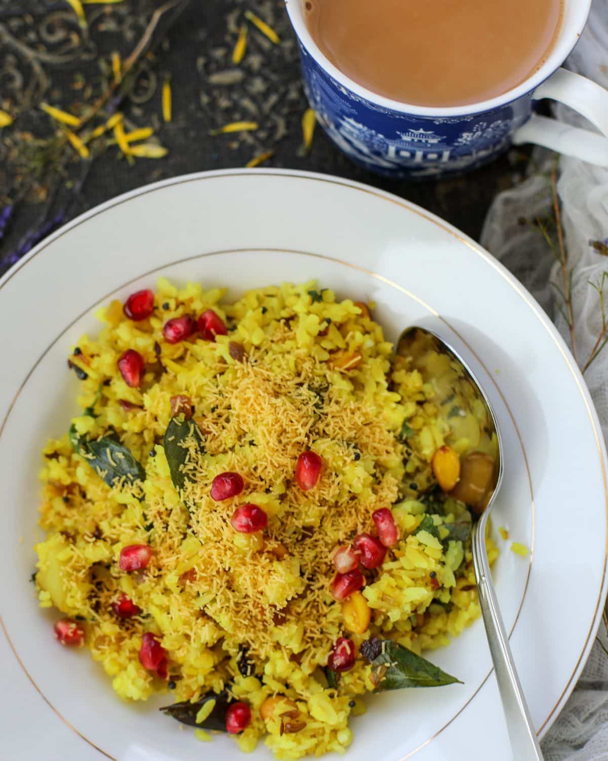 A plate of yellow poha garnished with sev, curry leaves, and pomegranate seeds, served with a spoon; a cup of chai is placed nearby.