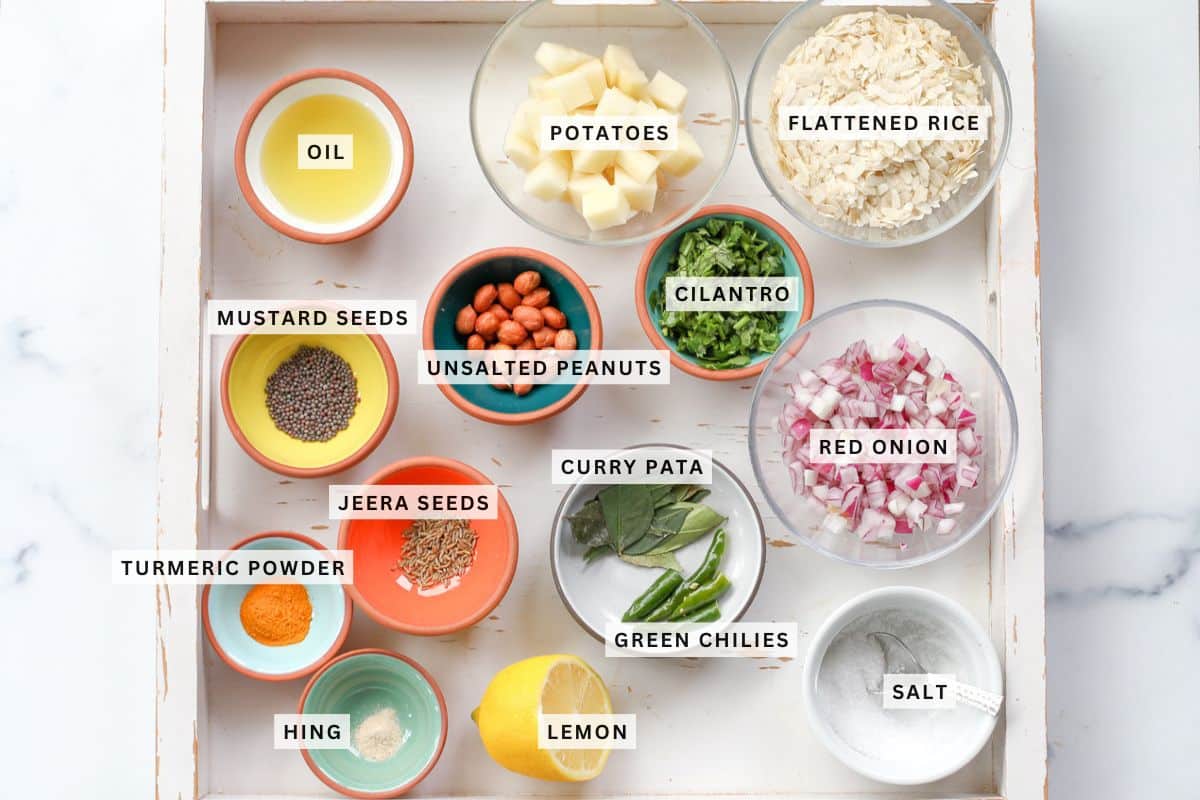 Tray with bowls containing oil, diced potatoes, flattened rice, cilantro, unsalted peanuts, red onion, mustard seeds, jeera seeds, curry pata, turmeric powder, hing, green chilies, lemon, and salt.