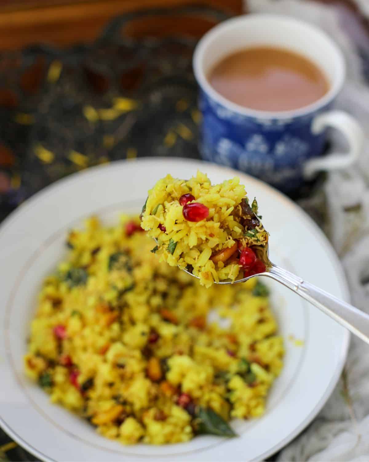A close-up of a spoonful of yellow rice with pomegranate seeds, held above a plate of the same dish, with a cup of tea in the background.