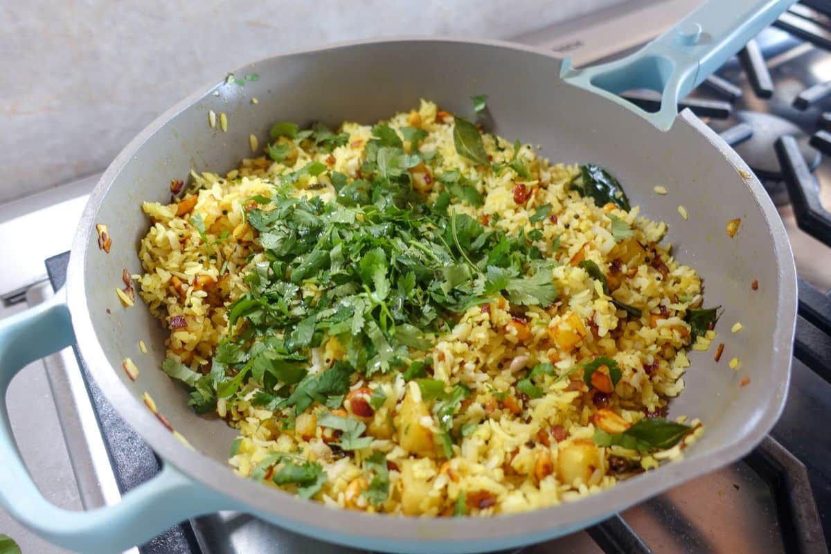 A pan on a stovetop filled with aloo poha, diced vegetables, and garnished with fresh cilantro.