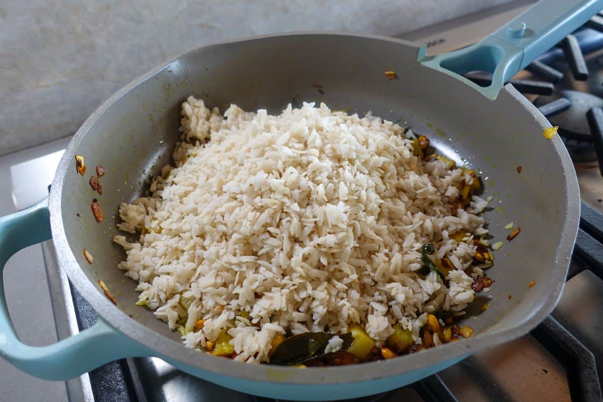 Cooked flattened rice added to a pan with sautéed vegetables and spices, sitting on a stove, ready to be mixed.