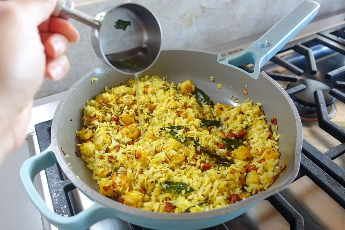 A hand pours liquid from a small metal cup into a pan of aloo poha with potatoes and spices on a stovetop.